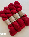 Three skeins of vibrant magenta-red yarn with paper bands, arranged on a light off-white background. The COLOR name 'BOBBIN'' .