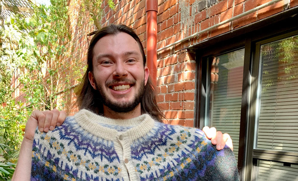 A young man with long dark hair is holding a fair isle sweater