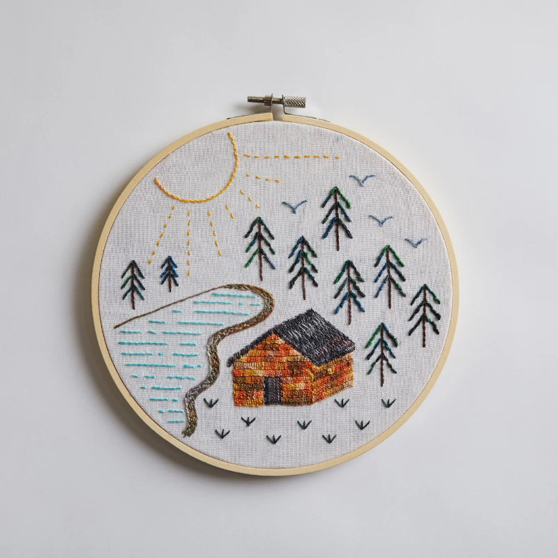 Embroidery hoop with a landscape design featuring a cabin, trees, and a sun on a light gray background.