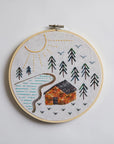 Embroidery hoop with a landscape design featuring a cabin, trees, and a sun on a light gray background.