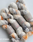 Four skeins of variegated yarn arranged on a light gray background. The yarn is primarily off-white/cream with orange, dark gray, and black variegated streaks.