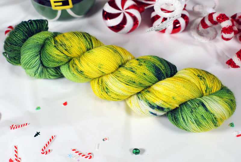 Two skeins of sparkly green and yellow yarn on a white background with Christmas-themed decorations.